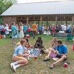 People sitting on the grass eating in the foreground with more people and a pavilion in the background