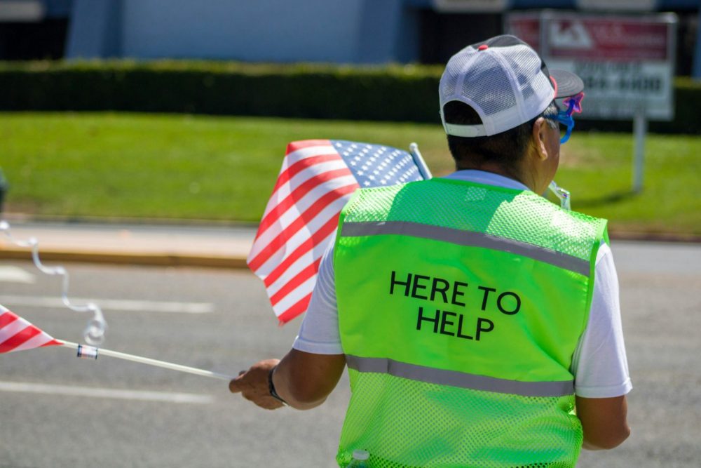 Man holding an American flag wearing a vest saying "here to help"
